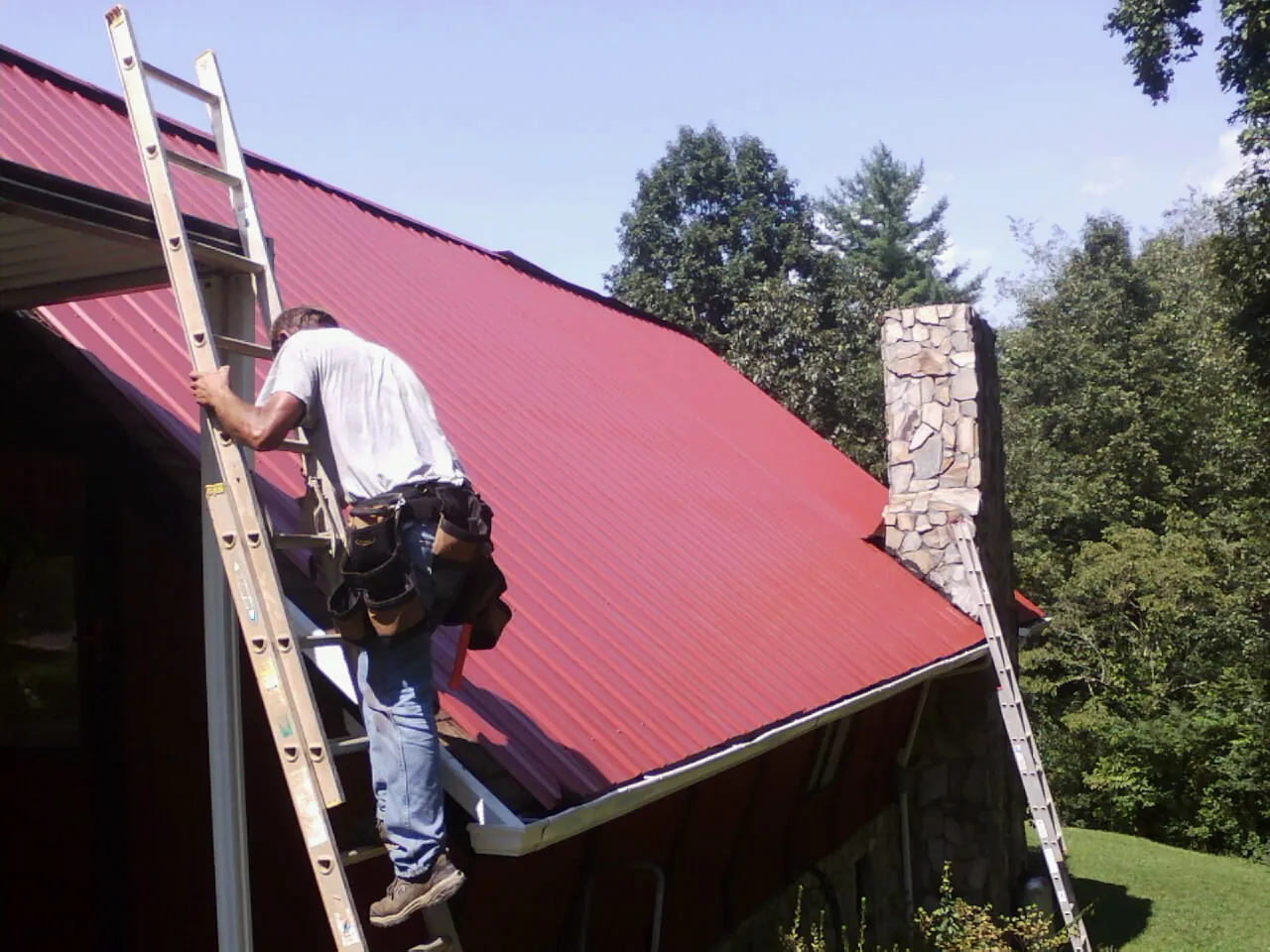 Roofer on a ladder beside a red metal roof and stone chimney — Soffit & Fascia Repair by Roofing Brothers in Elbing